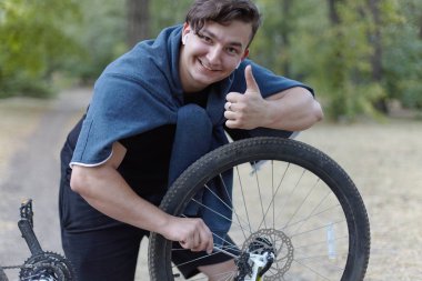 Koyu saçlı beyaz delikanlı tornavida ile terk edilmiş park yere serme Bisiklet ile çalışır. İş ile eğlenceli kavramı, başparmak, beyaz kulaklık; gülümseyerek. Açık havada, kopya alanı. 