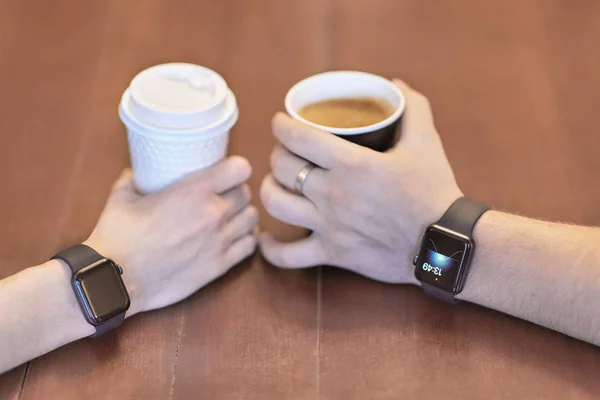 Two hands, male and female, both with equal electronic wrist watches, holding cups of coffee, white and black, on the wooden table. Date or friends meeting. Indoors, copy space.