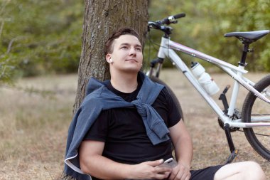 Mona Lisa gülüşü olan yakışıklı beyaz adam cep telefonu yere ağaç yakın terk edilmiş Parkı'nda oturan tutar. Bisiklet, kulaklık, raslantısal spor giyim. Açık havada, romantik bak, yalnızlık