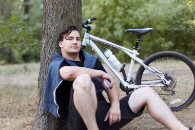 Genç yakışıklı beyaz adam üstünde hareket eden telefon, büyük ağaç yakın yerde terk edilmiş parkta oturan selfie yapar. Bisiklet arka plan üzerinde. Beyaz radyo earphones, mavi rahat spor giyim. Açık havada, spor aktivite sonrası dinlenmek.