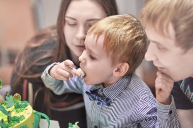 Beyaz sarışın yakışıklı tatlı gelen veliler arasında oturan doğum günü pastası yiyor. Mutlu doğum günü ve ebeveynlik kavramı. Kapalı, seçici odak, kopya alanı.