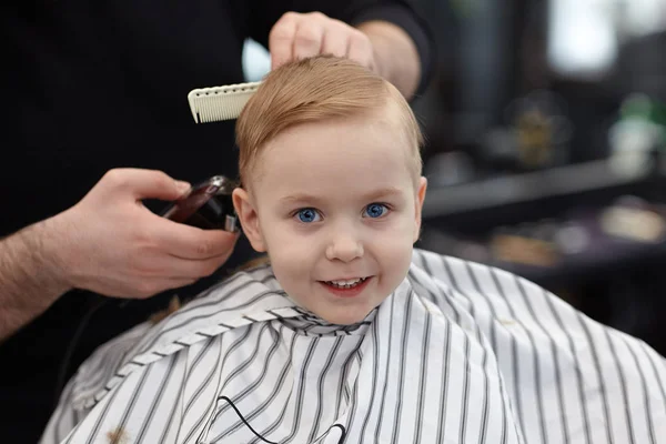 Sevimli sarışın bebek boy gülümseyerek Kuaför tarafından saç kesimi olan bir kuaför dükkanında mavi gözlü. Stilist araçları ile elleri. Çocuk moda. Kapalı, karanlık arka plan, kopya alanı.