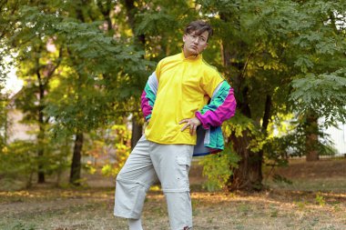 70 'lerin tarzında parlak spor kıyafetleri olan yakışıklı beyaz bir adam bir sabah parkında duruyor. Ağzına sopa, altın gözlük, küpe ve yüzük. Müsriflik modası. Açık hava, boşluğu kopyala.