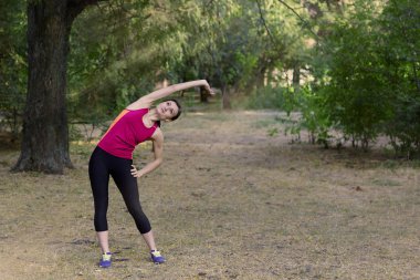 Muhteşem genç ince kadın sabah parkı spor salonu yapar. Yan eğim, parlak spor giyim, beyaz kulaklıklar, sakinlik ve dinlenme atmosferi, esneme eğitimi. Açık havada, kopyalama alanı.