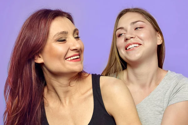 Two young laughing women, one caucasian blonde, another one latin. Beautiful diversity, fun and tight relationship, strong emotions. Violet background, indoors, copy space.