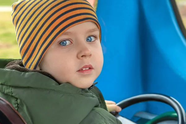 Bir oyun alanında elektrikli aracın direksiyonuna parlak mavi gözleri ile küçük güzel kafkas erkek bebek. Sürüş eğitimi, merak bakışı. Bahar kıyafeti, çizgili şapka. Açık havada, kopyalama alanı.
