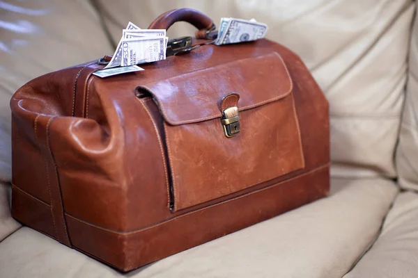 Old vintage red leather case full of money coming out of the sides. Scratched bag on leather sofa, business, bribe or prize concept. Indoors, copy space.