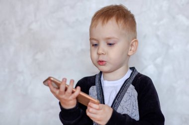 Mavi gözlü sevimli küçük sarışın erkek bebek cep telefonu aşağı bakar ve bir şey söylüyor. Çocuklar ve modern teknoloji konsepti. İç mekanlar, ışık arka planı, seçici odaklama, kopyalama alanı.