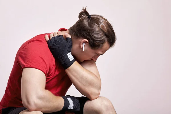 Young caucasian man with white earphones and little pony tail, in sportswear and boxing bandages sits in depressed pose. Despair moment, sorrow, hopelessness, despond before or after match. Indoors, copy space, light  background.