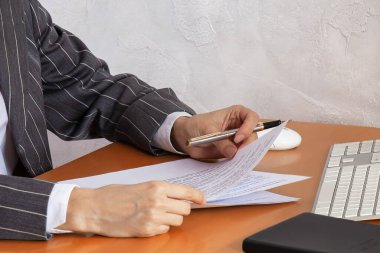 Gri çizgili takım elbiseli, elinde pahalı kalem ve kağıt belgeler tutan iş kadınının ellerini kapat. Beyaz klavye ve bilgisayar faresi, siyah defter ahşap masada yatıyor, açık arkaplan duvarı. 