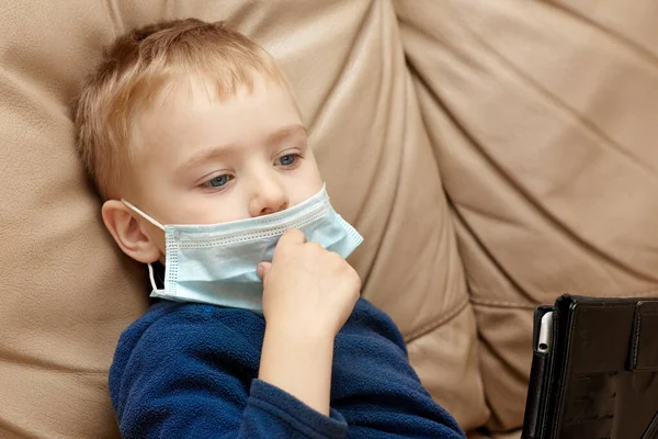 Sarı saçlı, mavi gözlü, sağlık maskeli, bej deri koltukta dijital tabletle oturan tatlı bir bebek. Mavi pijama, hastalık ya da virüs konsepti, kapalı alanda, kopyalama alanı.