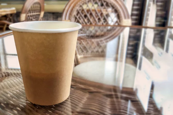 Close up craft cardboard cup with coffee on glass and straw table. Wooden chairs, interior of a small cafe in warm colors. Ecological biodegradable food grade tableware. Conscious consumption.