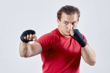 Genç adam boks yapıyor, sağ elle kanca atıyor. Siyah bandajlar, kırmızı tişört, kararlı yüz ifadesi, güçlü duygular. Öldürmek ya da vurmak için darbe kavramı, ışık arkaplanı üzerine boşluk kopyala.