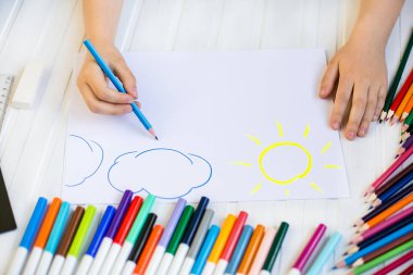 Çocuğun elleri ahşap bir masa üzerinde beyaz kağıda renkli kalemler ile boyanır.