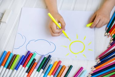 Çocuğun elleri ahşap bir masa üzerinde beyaz kağıda renkli kalemler ile boyanır.