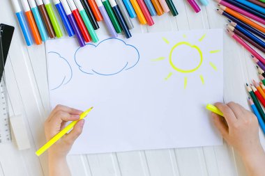 Çocuğun elleri ahşap bir masa üzerinde beyaz kağıda renkli kalemler ile boyanır.