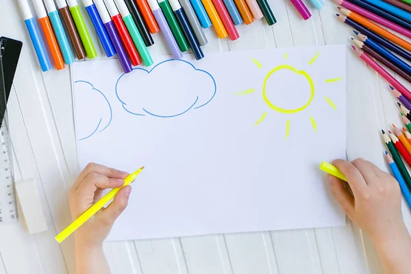 Çocuğun elleri ahşap bir masa üzerinde beyaz kağıda renkli kalemler ile boyanır.