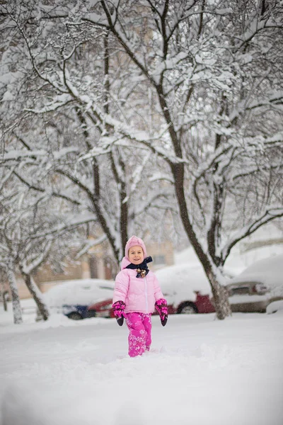 The child plays with snow in the winter. A little girl in a bright jacket and knitted hat, catches snowflakes in a winter park for Christmas. Children play and jump in the snow-covered garden.