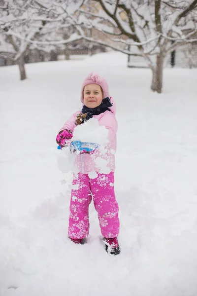 The child plays with snow in the winter. A little girl in a bright jacket and knitted hat, catches snowflakes in a winter park for Christmas. Children play and jump in the snow-covered garden.