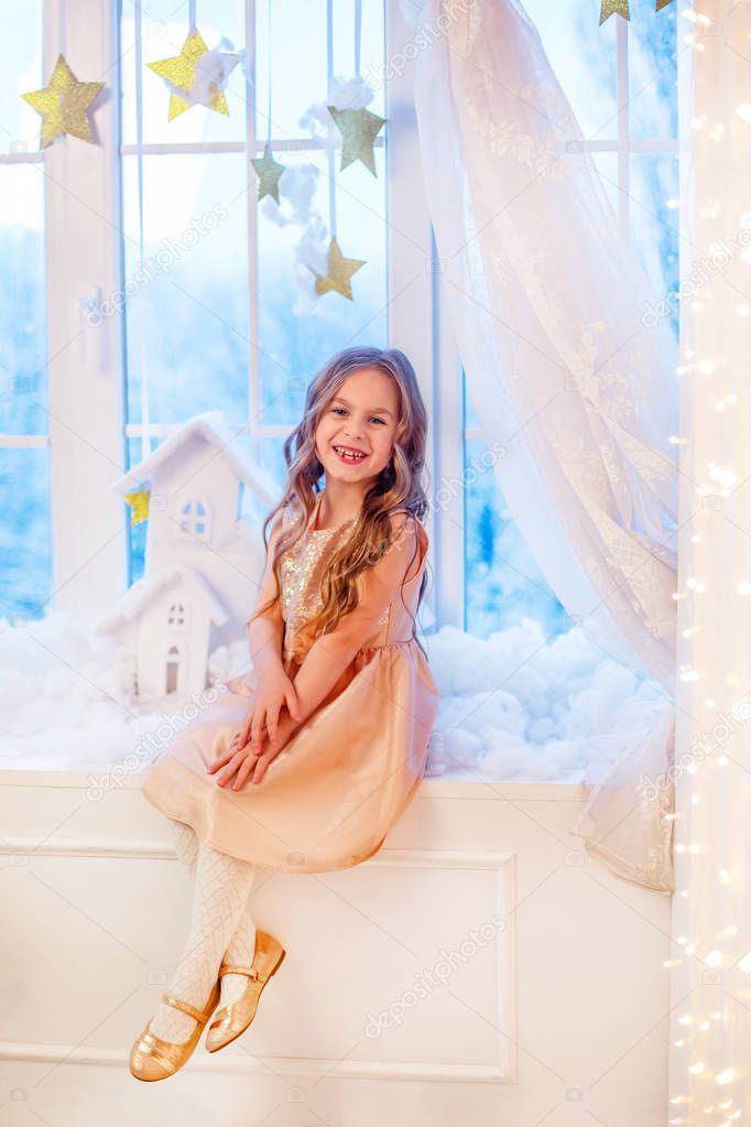 Cute little girl with curly hair at the window in anticipation of Christmas and New Year's magic. Funny emotions in winter day.