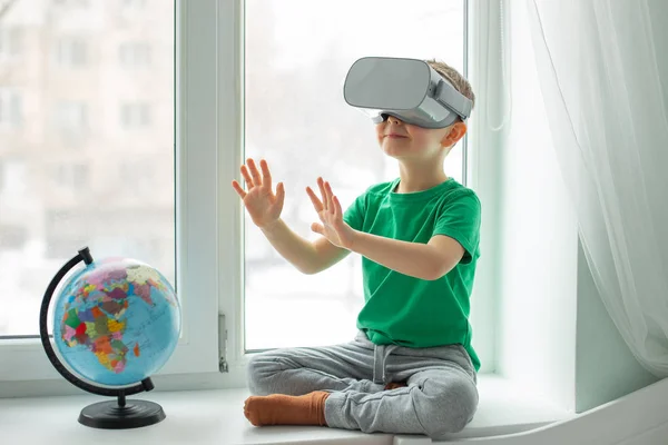 A boy with a virtual reality headset is sitting at home at the table indoors. The child explores the world of virtual reality through video and games. Technologies of the future.
