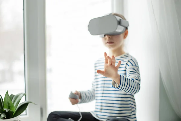 A child with a virtual reality headset is sitting at the table indoors at home. A girl exploring the world of virtual reality through video and games. Technologies of the future.