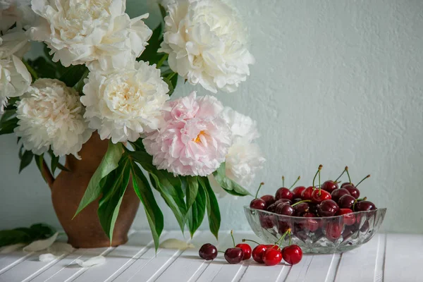 A large bouquet of peonies in a ceramic vase on the table, cherries in a bowl. Romantic mood.