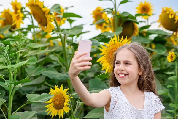 Chica hace selfie en un teléfono inteligente con una sonrisa en su cara, sobre el fondo de un campo de girasoles . —  Fotos de Stock