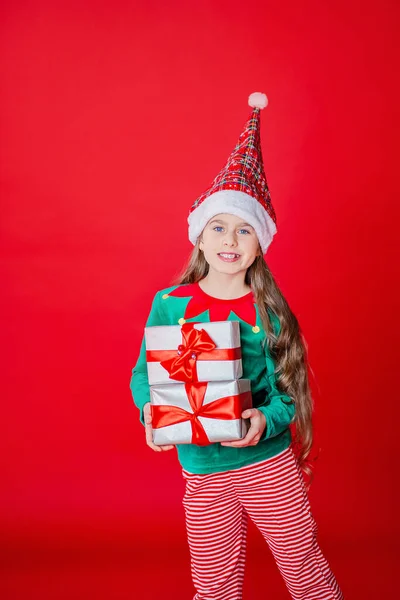 Merry Christmas, happy attractive girl with gifts in a costume of Santa Claus helper elf on a bright red bright color background. Portrait of a beautiful elven baby. Copy space.