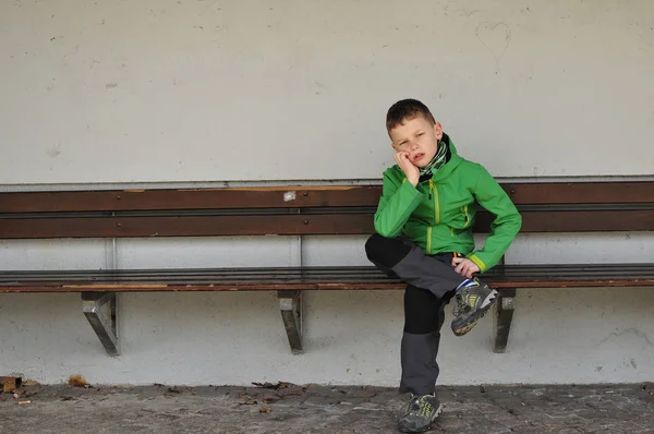 Um menino com uma jaqueta verde senta-se em uma parada de ônibus em um banco sozinho e espera o ônibus com espaço livre — Fotografia de Stock
