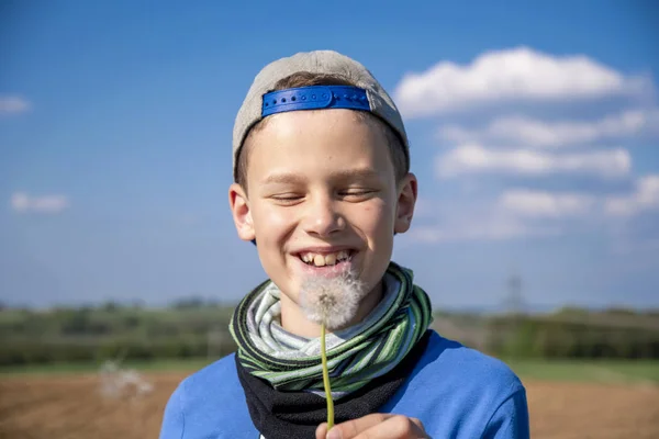 Boy with shield cap blows a flowering dandelion in front of a blue sky — Stock Photo, Image