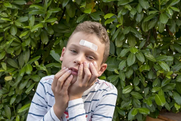 Menino com gesso no rosto na frente de uma sebe parece triste — Fotografia de Stock