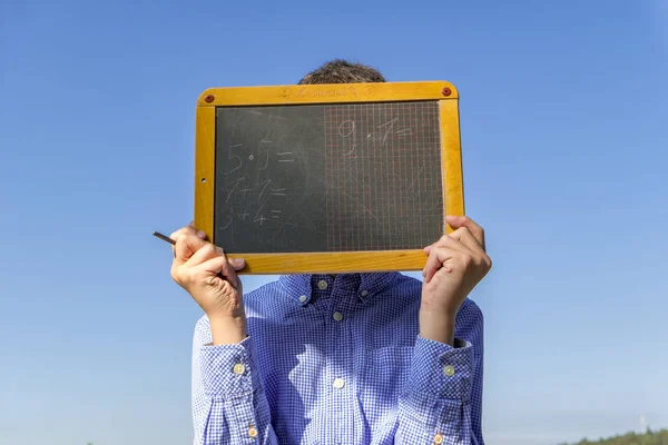 Menino segurando um quadro negro com tarefas aritméticas diretamente na frente de seu rosto — Fotografia de Stock