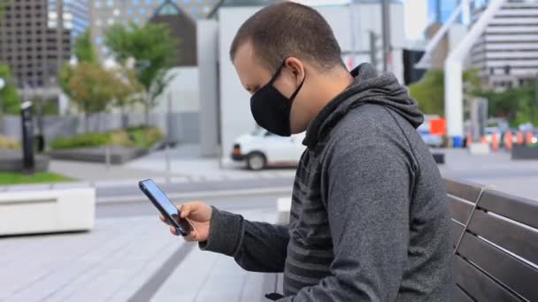 Mann mit Maske sitzt mit Handy auf Bank — Stockvideo