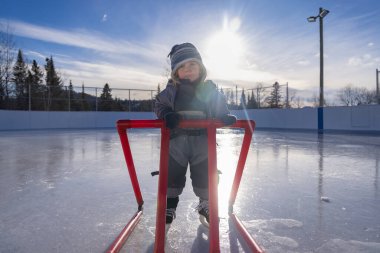 Young child learning to ice skate using a red support frame on a sunny winter day at an outdoor ice rink