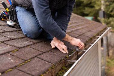 Construction worker tightening screws on gutter system using screwdriver while kneeling on rooftop of house