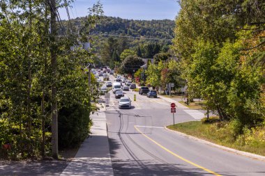 Traffic entering a picturesque town on a sunny day, surrounded by lush green trees and hills, creating a peaceful and inviting atmosphere