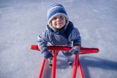 Happy little kid learning to ice skate using a red support frame on a sunny winter day at an outdoor ice rink, enjoying healthy activity