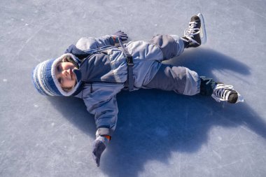 Kış kıyafetleri ve buz pateni giymiş küçük bir çocuk, donmuş bir buz pateni pistinde yatıyor, kış eğlencesi ve eğlencenin tadını çıkarıyor.