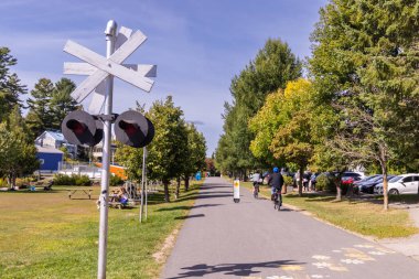 Bisikletçiler güneşli bir günün tadını çıkarıyorlar. Demiryolu kavşağının yanındaki kaldırımlı yolda ilerliyorlar. Ağaçlar ve çimenler bölgeyi kuşatıyor.