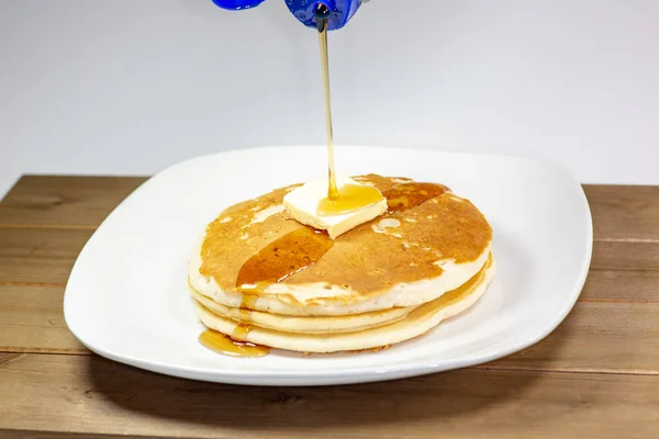 Pouring syrup from a blue tip bottle onto the square of butter on a stack of golden pancakes waiting to be eaten