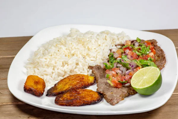 A steak covered in pico de gallo surrounded by plantains and white rice on a white plate. Cuban food