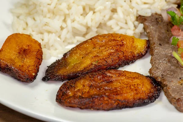 A steak covered in pico de gallo surrounded by plantains and white rice on a white plate. Cuban food