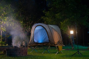 Çimlerin üzerinde kamp çadırı ve gaz lambası ormandaki bir odun sobasının yanında..