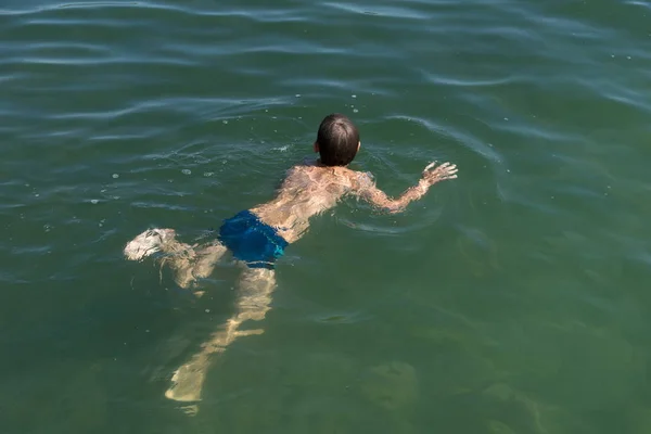 A boy of 11 years old is learning to swim in the blue water of a natural pond.
