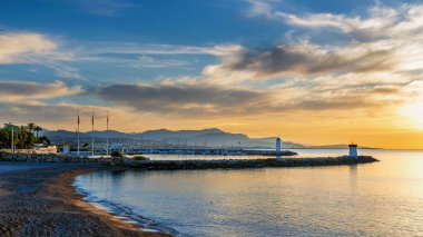 Deniz fenerlerinin arka planında deniz üzerinde gün doğumu, yatlı koylar, dağlar, Fransız Rivierası'nda bulutlarla gökyüzü