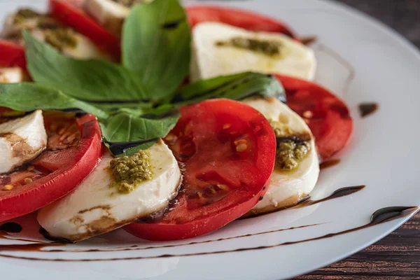 Assiette blanche de salade de caprese classique saine avec tomates et fromage mozzarella avec des feuilles de basilic et des lanières de sauce soja sur le fond en bois. Gros plan . — Photo