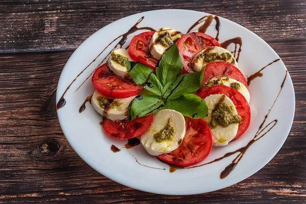 Assiette blanche de salade de caprese classique saine avec tomates et fromage mozzarella avec des feuilles de basilic et des lanières de sauce soja sur le fond en bois. Concept italien . — Photo