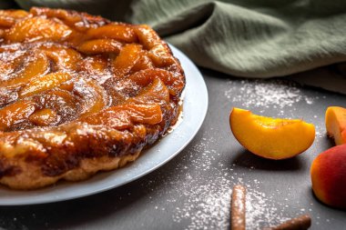 Şeftali, karamel ve pudra şekeri ile Tarte Tatin keten peçete ile karanlık bir arka plan üzerinde beyaz bir plaka üzerinde yakın çekim. Şeftali ve tarçın çubuklarının yanında. Yatay yönlendirme
