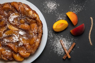 Tarte Tatin şeftali ile, karamel ve pudra şekeri koyu bir arka plan üzerinde beyaz bir plaka üzerinde. Şeftali ve tarçın çubuklarının yanında. Üst görünüm, yatay yönlendirme